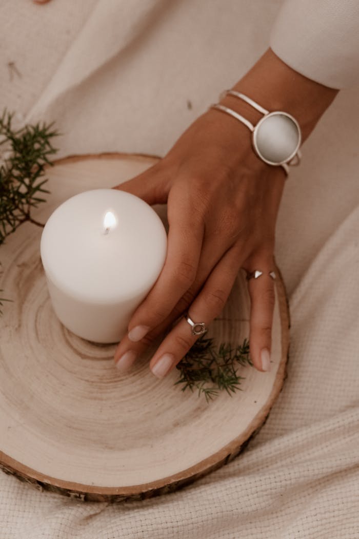 A woman's hand with jewelry holds a burning candle on a wood slice indoors, creating a cozy ambiance.